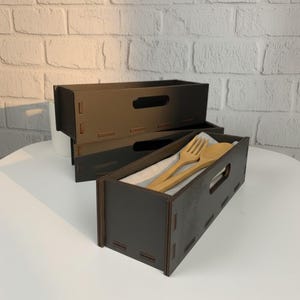 May include: Dark brown wooden utensil holders with a rectangular shape and a handle cutout. One holder contains a wooden fork and spoon on a linen cloth. The holders are stacked on a white surface.