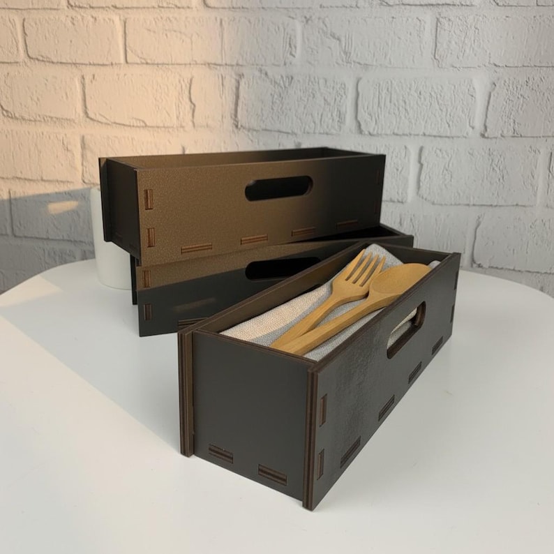 May include: Dark brown wooden utensil holders with a rectangular shape and a handle cutout. One holder contains a wooden fork and spoon on a linen cloth. The holders are stacked on a white surface.