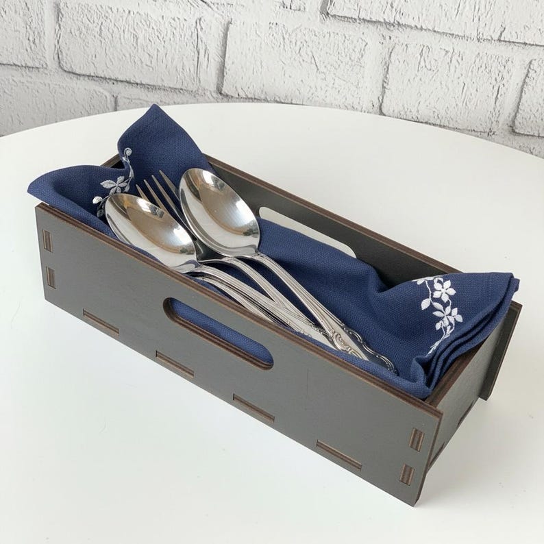 May include: A dark brown wooden cutlery holder with a navy blue napkin and silverware. The holder has a rectangular shape with cut-out handles. The napkin has a white floral design and holds two spoons and a fork.