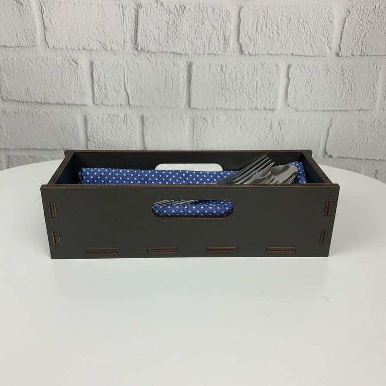 May include: A dark brown wooden cutlery holder with a rectangular shape and a handle cutout. Inside, a blue cloth with white polka dots holds silverware. The holder sits on a white surface against a white brick wall.