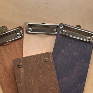 May include: A collection of four wooden clipboards featuring varying wood grain finishes in light, medium, and dark brown tones. Each clipboard is equipped with a polished silver metal clip mechanism at the top, secured with visible screws.