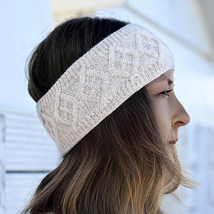 May include: A white knitted headband with a cable knit pattern. The headband is worn by a person with long brown hair.