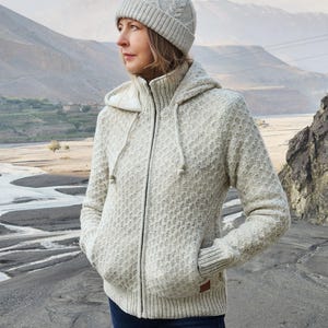 May include: A light gray hooded zip-up sweater and matching knit cap. The sweater has a textured pattern, a drawstring hood, and front pockets. The cap features a cable knit design. The outfit is suitable for outdoor activities.