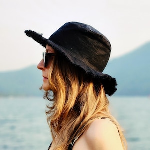 May include: A woman wearing a black sun hat with a frayed brim and sunglasses, standing near a body of water. She is wearing a black and white striped dress. The background features a blurred view of the water and mountains.