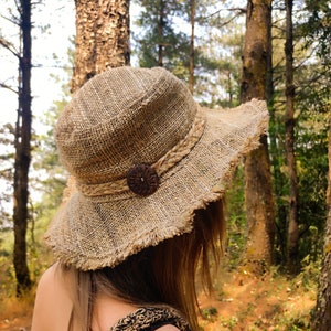 May include: A brown, woven, wide-brimmed sun hat with a frayed edge and a brown button detail. The hat is worn by a person in a forest setting.