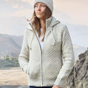 May include: A light gray knit zip-up hooded jacket with a matching beanie. The jacket has a textured pattern and a drawstring hood. The person is wearing blue jeans. The background features mountains and a river.
