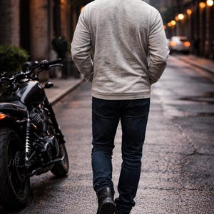 Op de afbeelding: Een persoon in een grijze hoodie, donkerblauwe jeans en zwarte laarzen loopt over een natte straat. Een zwarte motorfiets staat aan de kant van de weg. De sc&egrave;ne wordt verlicht door straatverlichting.