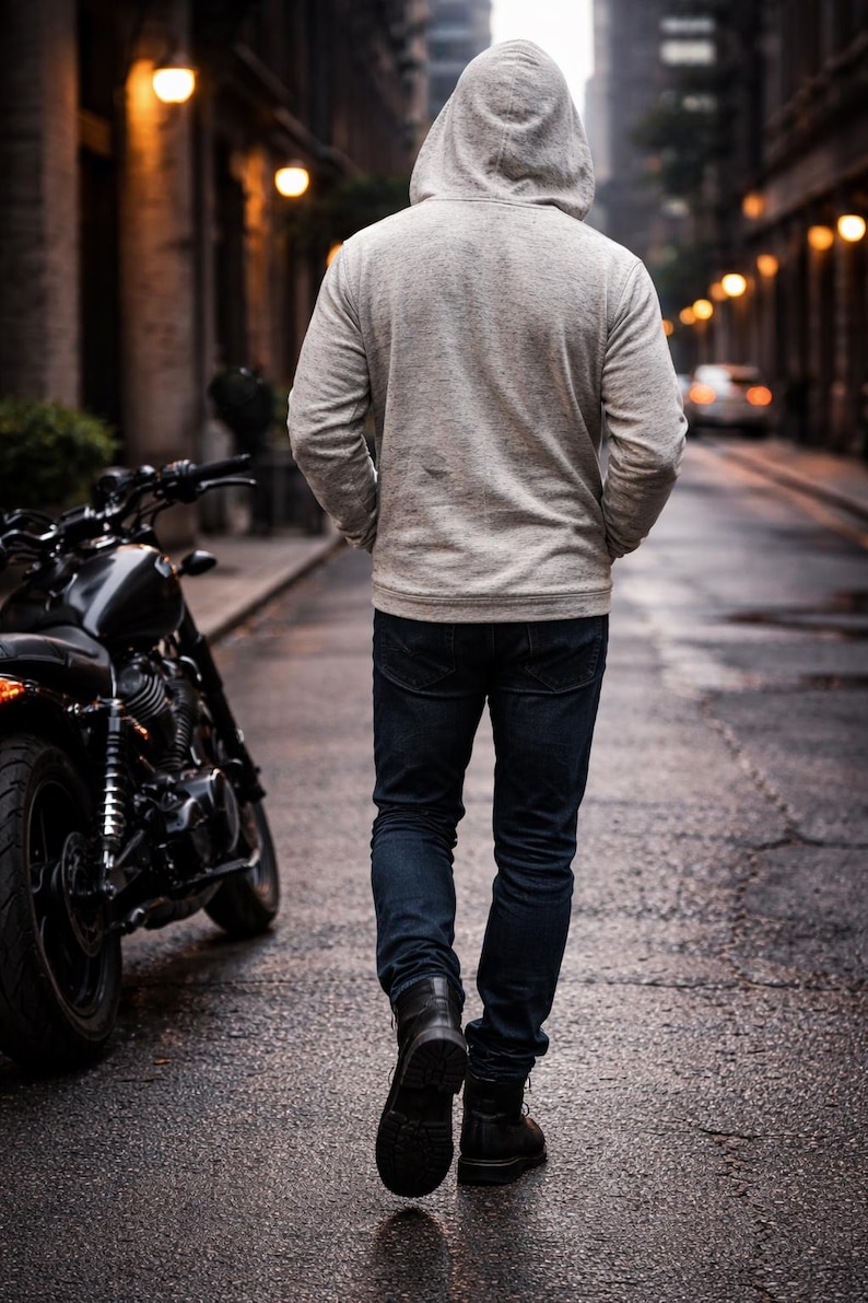 Op de afbeelding: Een persoon in een grijze hoodie, donkerblauwe jeans en zwarte laarzen loopt over een natte straat. Een zwarte motorfiets staat aan de kant van de weg. De sc&egrave;ne wordt verlicht door straatverlichting.