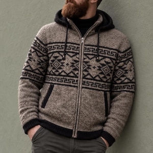 May include: A beige and black knit zip-up hoodie featuring a geometric pattern across the chest and sleeves. The garment includes a black hood, black ribbed cuffs, a black hem, and two front pockets with black trim, worn over a black turtleneck.