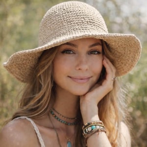 May include: A beige crocheted sun hat with a wide brim. The hat is made of a textured yarn and has a rounded crown. The hat is worn by a person with long blonde hair, turquoise jewelry, and multiple bracelets.