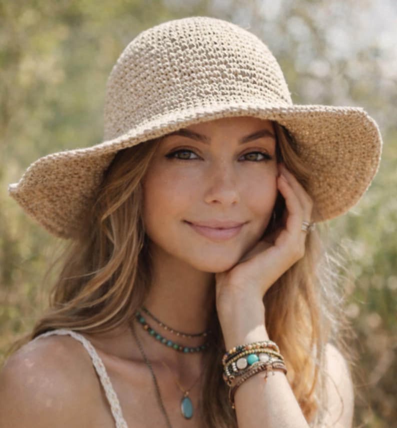 May include: A beige crocheted sun hat with a wide brim. The hat is made of a textured yarn and has a rounded crown. The hat is worn by a person with long blonde hair, turquoise jewelry, and multiple bracelets.