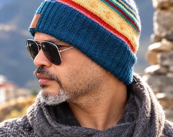 May include: A knit beanie featuring a wide, ribbed dark blue cuff and a crown patterned with horizontal stripes in yellow, red, and teal. A small, rectangular brown leather patch is attached to the side of the ribbed cuff, adding a subtle detail.