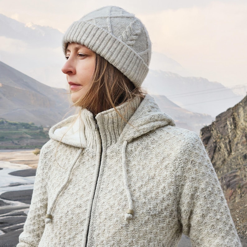 May include: A light gray knit hat with a leaf design and a matching zip-up hooded sweater. The hat has a ribbed brim and a textured pattern on the crown. The sweater has a textured pattern and a high collar.