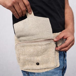 K&ouml;nnte beinhalten: Beige H&uuml;fttasche aus Hanf mit einem Rei&szlig;verschluss-Hauptfach und einer kleineren Vordertasche. Die Tasche hat einen Klappenverschluss und einen schwarzen Knopf. Die Person tr&auml;gt eine blaue Jeans und ein schwarzes T-Shirt.