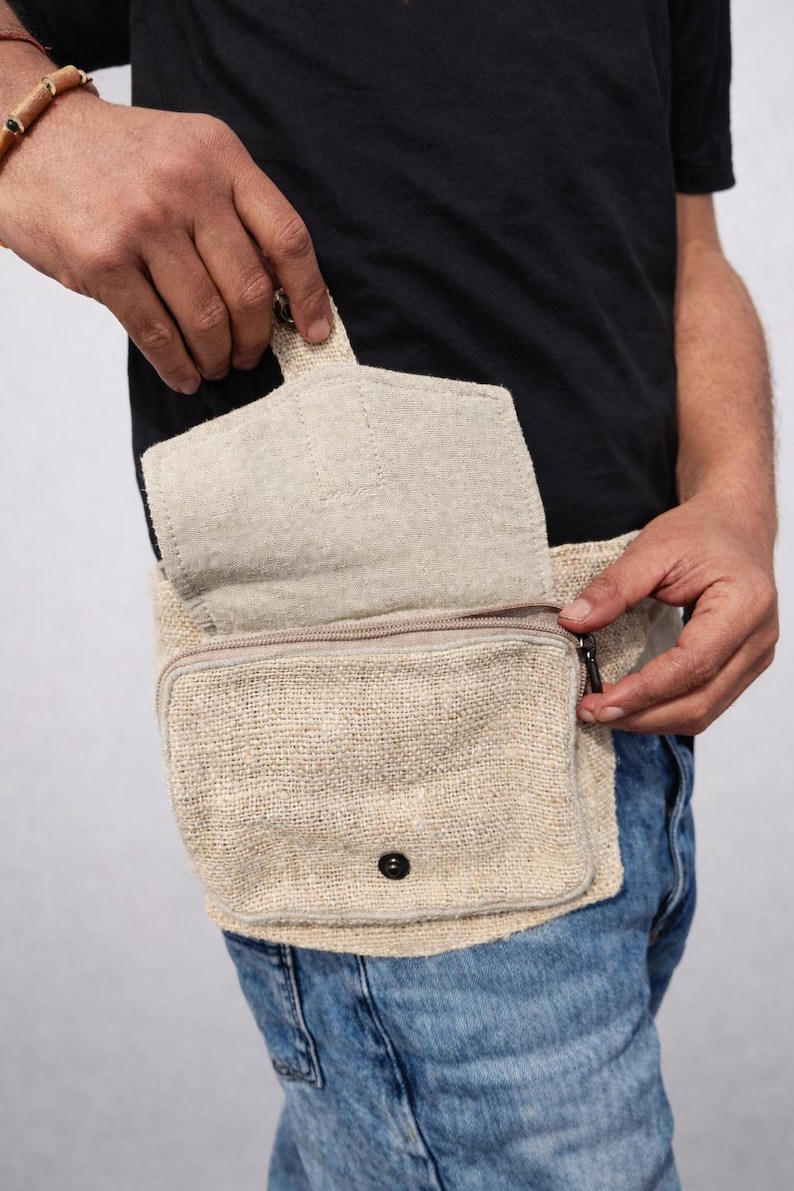 K&ouml;nnte beinhalten: Beige H&uuml;fttasche aus Hanf mit einem Rei&szlig;verschluss-Hauptfach und einer kleineren Vordertasche. Die Tasche hat einen Klappenverschluss und einen schwarzen Knopf. Die Person tr&auml;gt eine blaue Jeans und ein schwarzes T-Shirt.