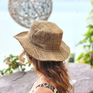 May include: A woven straw hat with a wide brim. The hat is a natural tan color and has a slightly textured appearance. The person is wearing a patterned top. The background includes a blurred decorative sphere and foliage.