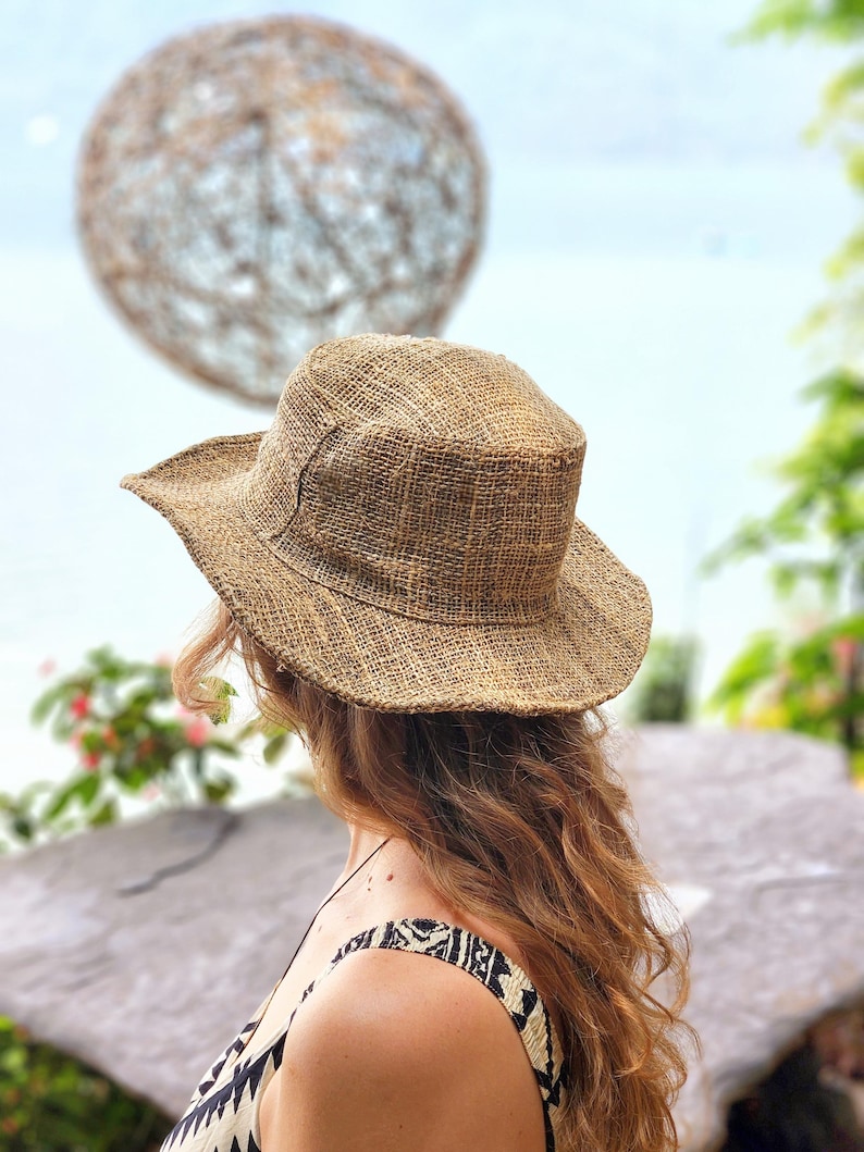 May include: A woven straw hat with a wide brim. The hat is a natural tan color and has a slightly textured appearance. The person is wearing a patterned top. The background includes a blurred decorative sphere and foliage.