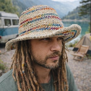 May include: A beige crochet hat with a colorful striped pattern. The hat has a wide brim and is worn by a person with long dreadlocks. The background includes a lake, mountains, and a van.