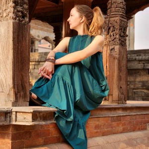 Pode incluir: Um vestido sem mangas verde-azulado com decote alto e bainha com babados. O vestido &eacute; usado por uma pessoa sentada em uma borda de tijolos. O fundo apresenta pilares de madeira esculpida e detalhes arquitet&ocirc;nicos.