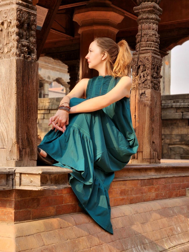 Pode incluir: Um vestido sem mangas verde-azulado com decote alto e bainha com babados. O vestido &eacute; usado por uma pessoa sentada em uma borda de tijolos. O fundo apresenta pilares de madeira esculpida e detalhes arquitet&ocirc;nicos.