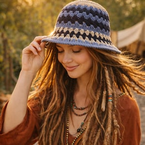 Op de afbeelding: Een gehaakte bucket hat in blauwe, zwarte en bruine tinten. De hoed heeft een patroon en een brede rand. De persoon op de foto draagt de hoed en een roestkleurige top. De achtergrond is wazig met warm, goudkleurig licht.