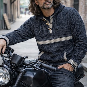 Op de afbeelding: Een donkergrijze hoodie met een cr&egrave;mekleurige horizontale streep over de borst en cr&egrave;mekleurige afwerking. De hoodie heeft houten knevelsluitingen en wordt gedragen met een donkere spijkerbroek. Een zwarte motorfiets staat op de voorgrond.