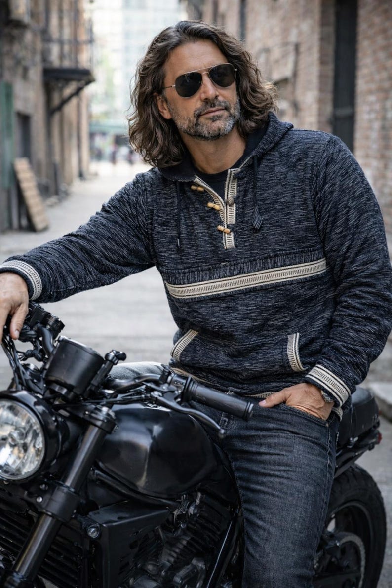 Op de afbeelding: Een donkergrijze hoodie met een cr&egrave;mekleurige horizontale streep over de borst en cr&egrave;mekleurige afwerking. De hoodie heeft houten knevelsluitingen en wordt gedragen met een donkere spijkerbroek. Een zwarte motorfiets staat op de voorgrond.