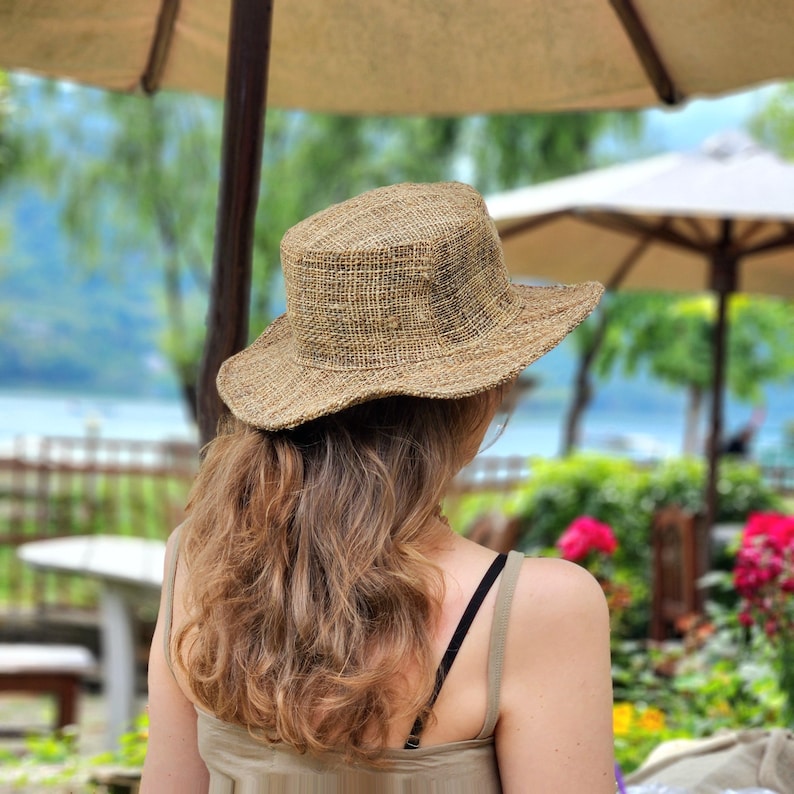 May include: A natural-toned straw hat with a wide brim. The hat has a slightly textured appearance and a round crown. The hat is worn by a person with long, wavy brown hair. The background includes a garden and a lake.