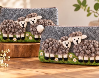 May include: Two rectangular felt pouches in gray, each featuring a textured applique of two sheep standing on a green grassy patch with white flowers. The pouches have a zipper closure with a small white felt ball attached to the pull.