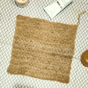 K&ouml;nnte beinhalten: Ein brauner, rechteckiger Bade-Schwamm aus gewebtem Material mit einem geknoteten Griff. Der Schwamm ist aus Naturfasern hergestellt und hat eine raue Textur.