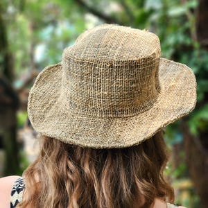 May include: A woven straw hat with a wide brim. The hat is a natural beige color and has a slightly textured appearance. The hat is designed to provide shade from the sun.