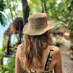 May include: A woven straw hat with a wide brim. The hat is a natural tan color and is worn by a person with long, wavy, light brown hair. The person is wearing a patterned dress, and the background is a blurred view of a lush, green outdoor setting.