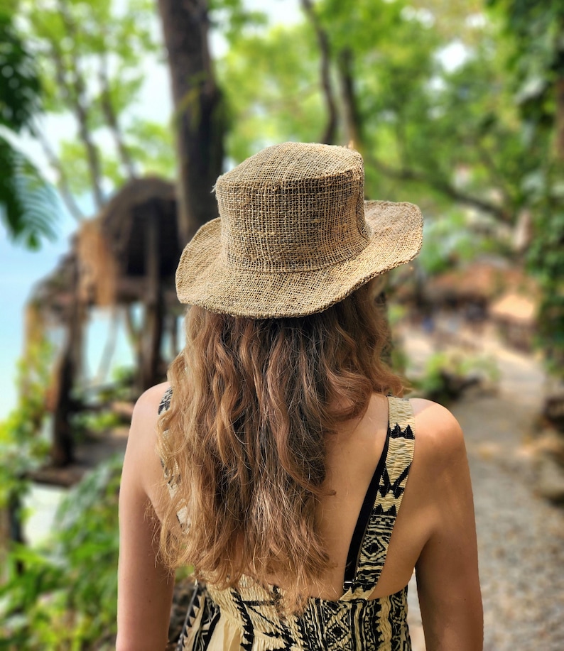 May include: A woven straw hat with a wide brim. The hat is a natural tan color and is worn by a person with long, wavy, light brown hair. The person is wearing a patterned dress, and the background is a blurred view of a lush, green outdoor setting.
