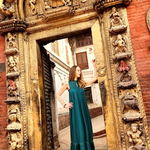 Pode incluir: Uma mulher em um vestido maxi verde-azulado est&aacute; em uma porta com detalhes ornamentados em madeira entalhada. A porta &eacute; emoldurada por tijolos vermelhos e o vestido tem uma bainha com babados. A moldura da porta &eacute; intrincadamente esculpida com figuras.