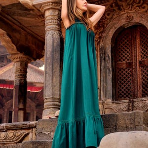 Pode incluir: Um vestido longo e esvoa&ccedil;ante verde-azulado com bainha com babados. O vestido tem um decote halter e &eacute; usado ao ar livre. O fundo apresenta arquitetura em pedra com detalhes esculpidos e uma porta de madeira.