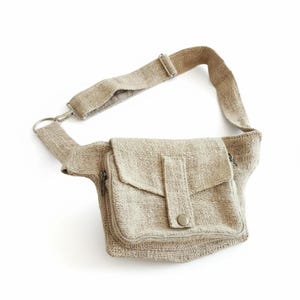 K&ouml;nnte beinhalten: Beige Bauchtasche aus gewebtem Stoff. Die Tasche hat ein Hauptfach mit Rei&szlig;verschluss, eine Vordertasche mit Klappe und Druckknopfverschluss sowie einen verstellbaren Riemen. Der Riemen ist mit einem Metallring an der Tasche befestigt.