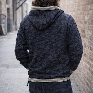 Op de afbeelding: Een donkerblauwe hoodie met een textuurgebreid patroon. De hoodie heeft een cr&egrave;me en beige gestreepte bies rond de capuchon, manchetten en taille. De persoon draagt een donkerblauwe jeans.