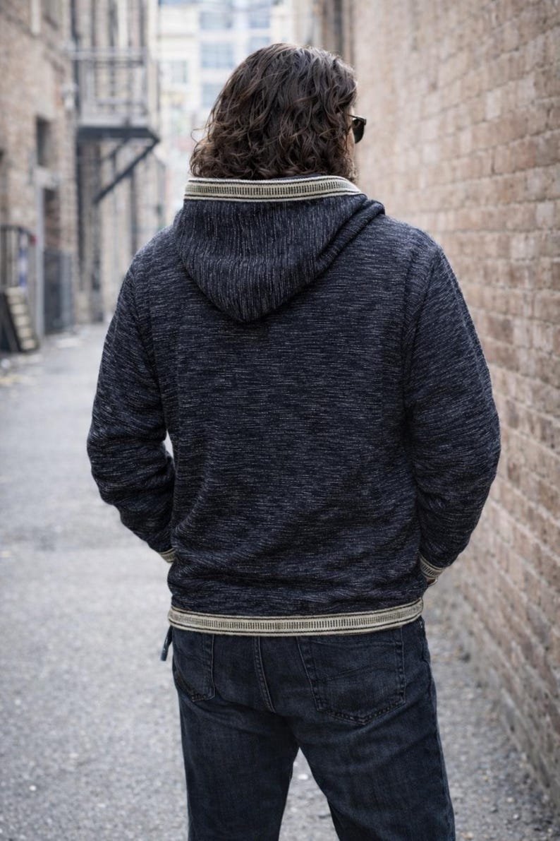 Op de afbeelding: Een donkerblauwe hoodie met een textuurgebreid patroon. De hoodie heeft een cr&egrave;me en beige gestreepte bies rond de capuchon, manchetten en taille. De persoon draagt een donkerblauwe jeans.