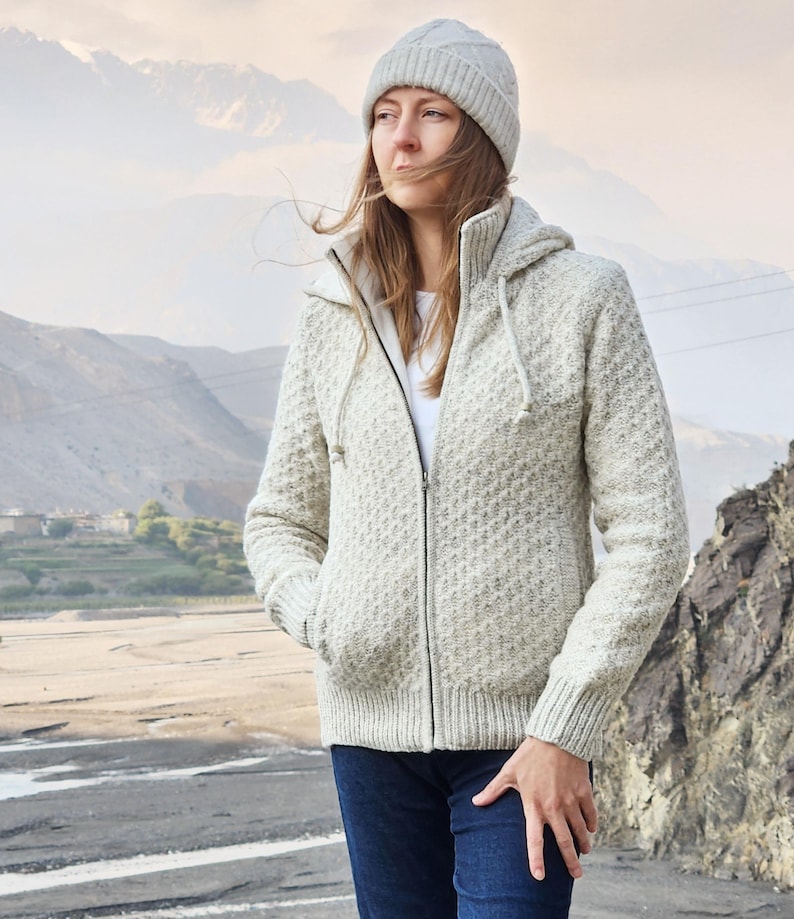 May include: A light gray knit zip-up hooded jacket with a matching beanie. The jacket has a textured pattern and a drawstring hood. The person is wearing blue jeans. The background features mountains and a river.