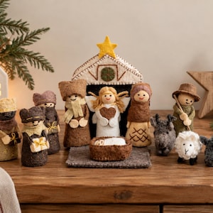 Pode incluir: Um conjunto de presépio em feltro com um estábulo com uma estrela amarela, um anjo, um bebé numa manjedoura e várias figuras, incluindo reis magos, um pastor e animais. As figuras são feitas de lã de feltro castanha, bege, branca e cinzenta.
