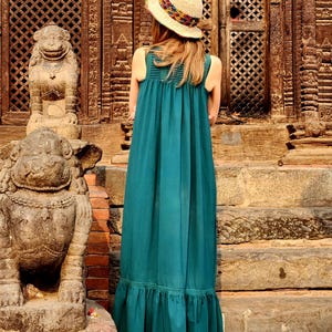 Pode incluir: Uma pessoa vestindo um vestido maxi verde-azulado e um chap&eacute;u de palha com uma faixa colorida, em p&eacute; em degraus de pedra. O vestido tem uma bainha com babados e um corpete franzido. O fundo apresenta portas de madeira esculpida e est&aacute;tuas de pedra.
