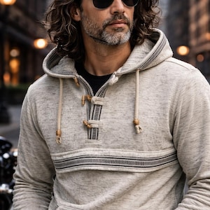 Op de afbeelding: Een lichtgrijze hoodie met een trekkoord en houten knevels. De sweatshirt heeft een horizontale streep over de borst en een voorzak. Het model draagt een zonnebril.