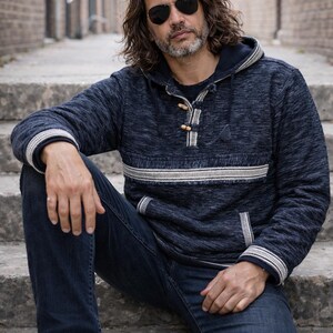 Op de afbeelding: Een marineblauwe hoodie met een witte streep en afwerking, gecombineerd met een donkerblauwe jeans. De hoodie heeft houten knevelsluitingen. De persoon draagt een zonnebril. De foto is buiten gemaakt.