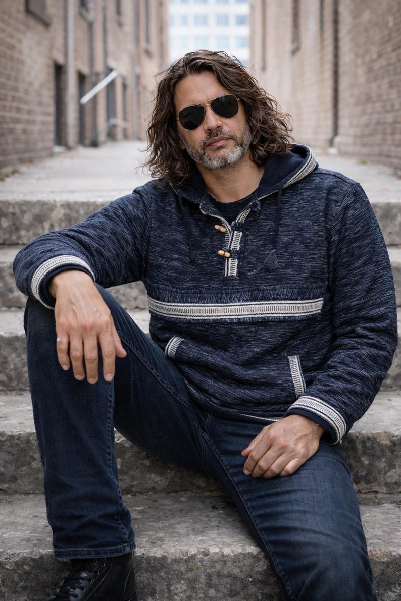 Op de afbeelding: Een marineblauwe hoodie met een witte streep en afwerking, gecombineerd met een donkerblauwe jeans. De hoodie heeft houten knevelsluitingen. De persoon draagt een zonnebril. De foto is buiten gemaakt.