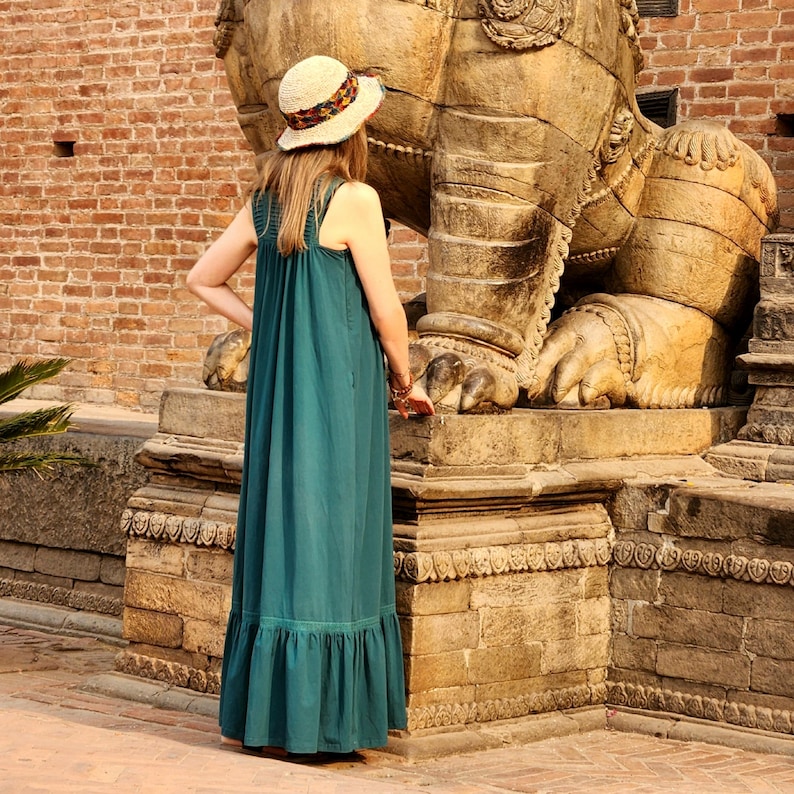 Pode incluir: Um vestido maxi verde-azulado com bainha com babados e um chap&eacute;u de palha com uma faixa estampada. O vestido tem um design simples e sem mangas. A pessoa est&aacute; em frente a uma grande escultura de pedra.