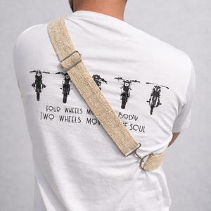 K&ouml;nnte beinhalten: Wei&szlig;es T-Shirt mit Motorrad-Grafik und dem Text "FOUR WHEELS MOVE THE BODY, TWO WHEELS MOVE THE SOUL." Ein beigefarbener, gewebter Gurt mit Metallbeschl&auml;gen wird &uuml;ber dem K&ouml;rper getragen. Der Gurt ist am T-Shirt befestigt.