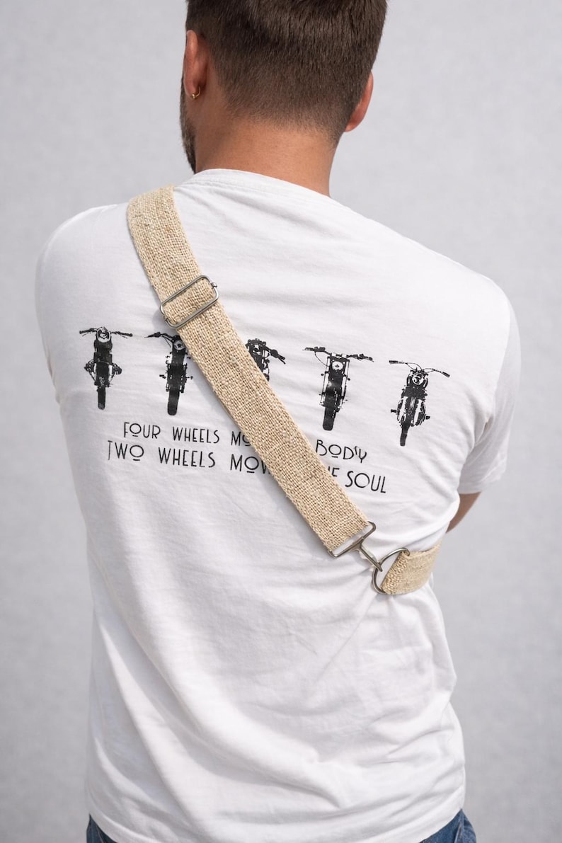 K&ouml;nnte beinhalten: Wei&szlig;es T-Shirt mit Motorrad-Grafik und dem Text "FOUR WHEELS MOVE THE BODY, TWO WHEELS MOVE THE SOUL." Ein beigefarbener, gewebter Gurt mit Metallbeschl&auml;gen wird &uuml;ber dem K&ouml;rper getragen. Der Gurt ist am T-Shirt befestigt.