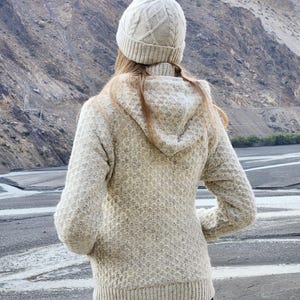 May include: A person wearing a light beige knit hat and matching hooded sweater. The sweater has a textured pattern and ribbed cuffs and hem. The person is wearing dark blue jeans and is standing outdoors with a mountain backdrop.