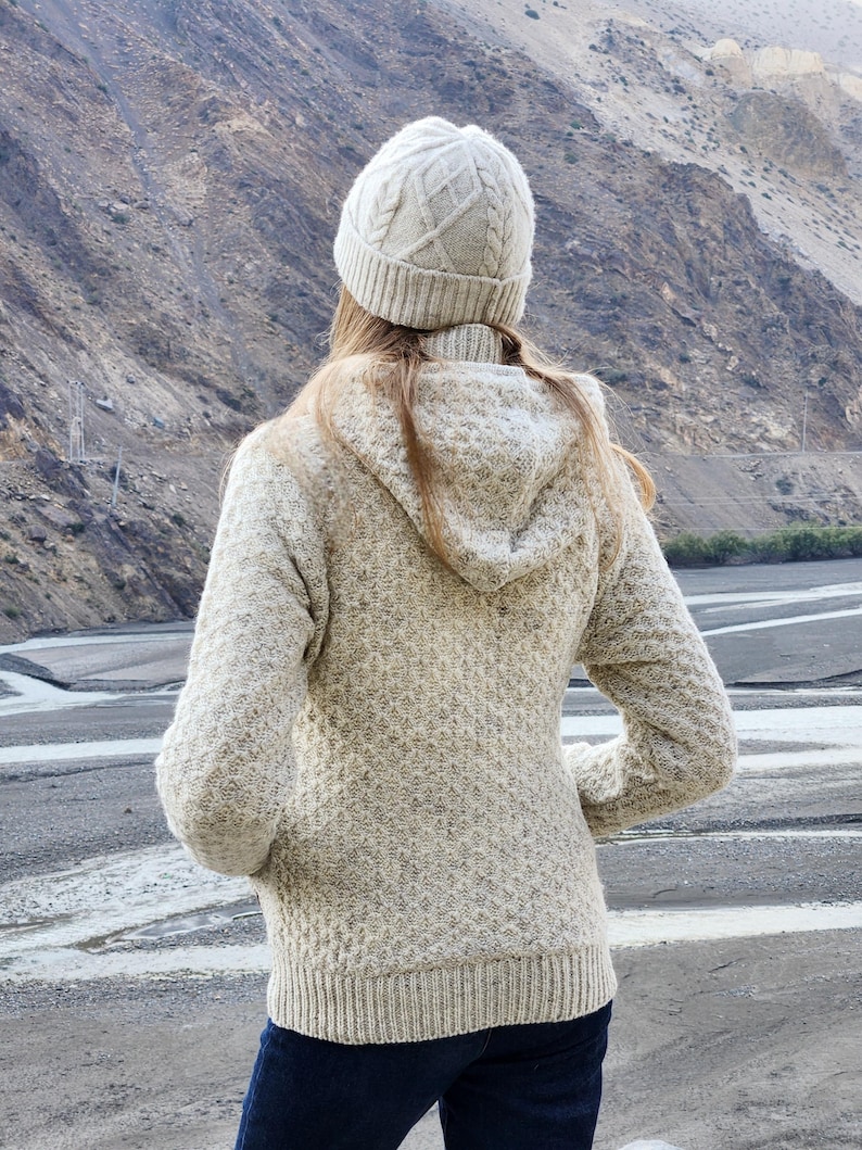 May include: A person wearing a light beige knit hat and matching hooded sweater. The sweater has a textured pattern and ribbed cuffs and hem. The person is wearing dark blue jeans and is standing outdoors with a mountain backdrop.