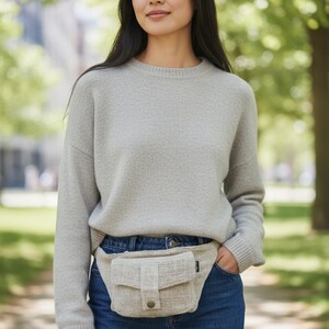 K&ouml;nnte beinhalten: Eine Frau tr&auml;gt einen hellgrauen Pullover und blaue Jeans mit einer beigen Bauchtasche. Die Bauchtasche hat eine rechteckige Form mit einer Klappe und einem Knopfverschluss. Der Hintergrund ist verschwommen mit B&auml;umen und einem Geb&auml;ude.